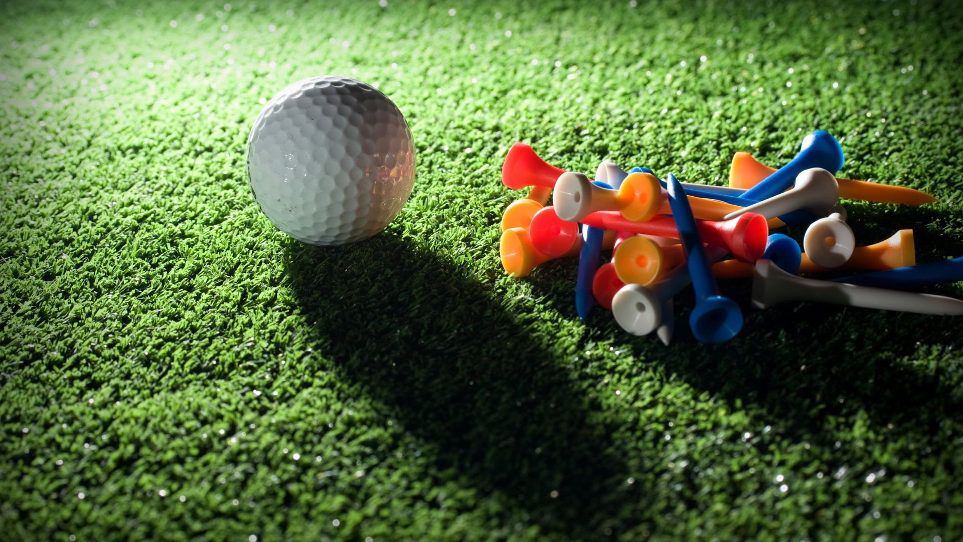A white golf ball sits on artificial turf next to a scattered pile of colorful plastic golf tees.